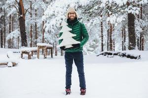 retrato de hombre con barba espesa y bigote tiene una expresión complacida, camina en un hermoso bosque de invierno, sostiene un pequeño abeto artificial blanco, lo anuncia antes de las vacaciones de año nuevo foto