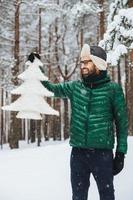 retrato vertical de un hombre barbudo positivo y encantador vestido con ropa cálida de invierno, mira con expresión feliz el abeto artificial, demuestra que protege la naturaleza y no tala árboles foto