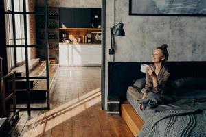 joven feliz y atractiva chica sentada en la cama en posición de loto y bebiendo café por la mañana foto