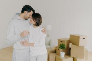 Lovely couple embrace and feel happy, holds keys from new modern first house, move in apartment for living together, pose in empty room with packed carton boxes, feel love to each other. Real estate photo