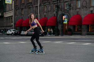 Pleased sporty woman rides on rollers in urban place leads healthy lifestyle dressed in sportsclothes looks away has cheerful expression enjoys fitness training. Sports activity hobby recreation photo