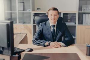 Proud handsome male entrepreneur sits in his cozy cabinet, works on computer, satisfied with successful working day, dressed formally, looks directly into camera. Intelligent employee in office photo