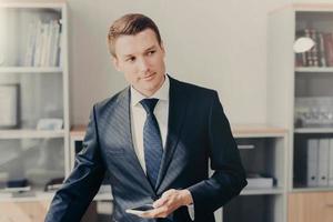 Horizontal shot of handsome male manager in stylish clothes, uses smart phone for checking information about entrepreneurship, reads news in web page, connected to wireless internet, stands at office photo