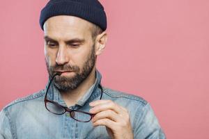 una foto de un hombre barbudo pensativo mira hacia abajo, sostiene anteojos, usa una chaqueta de mezclilla y un sombrero, aislado sobre un fondo rosado con espacio para copiar su contenido publicitario. personas y pensamientos