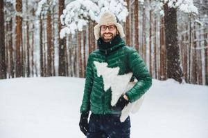 un alegre hombre barbudo usa un cálido anorak y un sombrero, camina en el bosque de invierno, sostiene un abeto artificial, tiene un ambiente festivo y va a celebrar el año nuevo. tiro al aire libre de macho pasa tiempo libre en clima frío foto