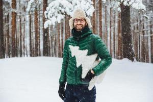 Cheerful bearded male wears warm anorak and hat, walks in winter forest, holds artificial fir tree, has festive mood, going to celebrate New Year. Outdoor shot of male spends free time on cold weather photo