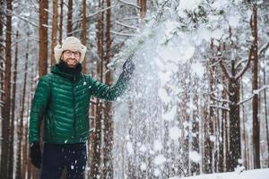 Portrait of cheerful handsome male wears fur cap with ear flap and yellow and green jacket, throws snow in air, enjoys free time spending in beautiful winter forest. People, relaxation concept photo