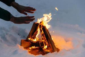 Unrecognizable male warms hands on fire in forest during cold winter, tries to warm himself, being cold spend time on cold snow and frosty weather. Burning flame on ground covered with snow. photo