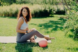 Positive brunette woman in cropped top and leggings poses bare feet on karemat uses ball for gymnastic exercises smiles pleasantly enjoys beautiful nature view. Sport outdoor. Yoga practice. photo