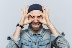 Portrait of hppy bearded male makes eyewear with fingers, has stubble, wears jeans cloting, stands against white background. Funny middle aged man foolishes indoor, pretends wearing glasses. photo