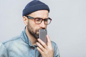 Thoughtful middle aged male with stubble holds smart phone near mouth, being deep in thoughts, thinks about future plans, isolated over white background. People, emotions, technology concept photo