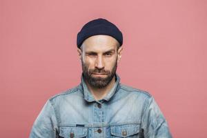 Photo of good looking bearded male blinks eye and looks seriously into camera, wears denim jacket and black hat, isolated over pink background. Stylish man with stubble poses in studio alone.