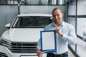 Smiling and having good mood. Manager stands in front of modern white car with paper and documents in hands photo