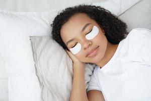 Young biracial girl with under eye patches on face relax lying in bed. Morning skincare routine photo