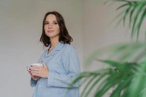 Photo of thoughtful young woman with dark hair, appealing appearance, warms with hot beverage, dressed in domestic costume, poses indoor near green plant, blank space for your advertisement.