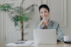 Charming stylish businesswoman works freelance at laptop computer, uses high speed internet connection, holds eyeglasses in hand, enjoys favorite remote job, reads publication, sits at office desk photo