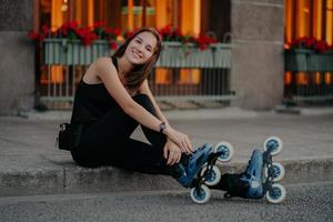 Una joven europea de aspecto agradable que usa patines se toma un descanso después de montar poses al aire libre vestida con ropa deportiva negra y tiene una sonrisa feliz en la cara. concepto de hobby y estilo de vida de recreación de personas foto