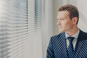 Prosperous male boss in elegant luxury suit, stands in modern office near big window with shutter, has rest after meeting with shareholders of company, thinks about something. People, work concept photo