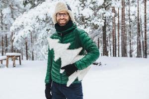 el hombre barbudo de aspecto agradable usa ropa abrigada, sostiene un abeto blanco, se para contra el fondo del bosque invernal, camina con amigos durante el clima helado, está de buen humor. gente y naturaleza foto