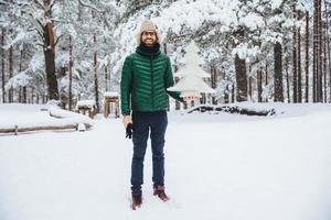 Outdoor shot of good looking male wears warm hat and green jacket holds winter fir tree, poses against trees covered with snow, looks happily directlly into camera. Recreation and season cpncept photo
