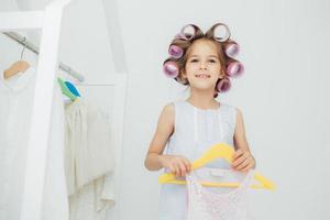 Indoor shot of fashionable cute small girl with gentle warm smile, prepares for something, has curlers on hair, holds new outfit on hangers, look joyfully at camera. Children and beauty concept photo