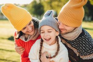 Adorable funny small kid has fun with parents who look at her with great love, enjoy spending free time together, admire beautiful nature and fresh air. People, happiness and childhood concept photo