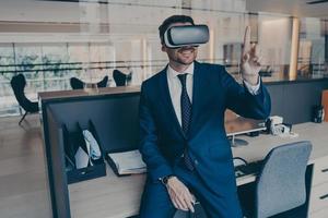 Businessman sitting on desk in office and using VR headset on his head, pointing with forefinger photo