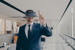 Smiling male entrepreneur using vr goggles for 3d visualization, pointing with finger photo