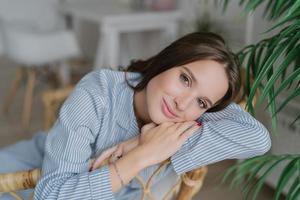 Image of good looking young female model with dark hair, sits on wooden chair, has relaxed facial expression, has charming smile, enjoys domestic calm atmosphere. Beauty and emotions concept photo
