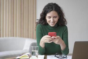 Young italian freelancer lady with dark wavy hair holding cell phone and looking at screen with smile photo