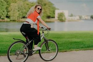 Image of female model rides bicycle, looks aside with cheerful expression, wears sunglasses, breathes fresh air, poses near lake and green trees, covers long distance, finds out something new photo