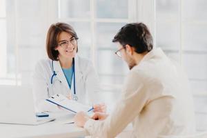 Woman doctor with stethoscope holds binder with patient personal medical card, consults patient who has medical problems, sit at hospital office, discuss medical checkup results, offers insurance photo