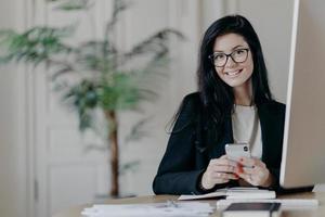 Happy female office worker in stylish black jacket browses smartphone with cheerful expression, sends text messages, works on important project, poses in coworking space. Modern technologies photo
