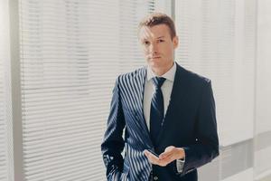 Prosperous male director sends message and multimedia files via smart phone, synchronizes phone data and shares photos for project, poses in office, wears luxury expensive suit, looks seriously
