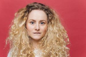 Headshot of beautiful curly blonde female with blue eyes, healthy skin, bites lower lip, looks directly into camera, isolated over bright pink background. People, appearance and beauty concept photo