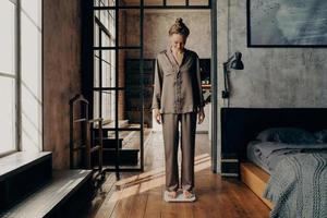 Young lovely female in comfy satin pajama standing on electronic scales in bedroom photo