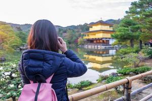 solo woman tourist taking photo by camera at Kinkakuji temple or the golden pavilion in Autumn season, Asian traveler visit in Kyoto, Japan. Vacation, destination and travel concept