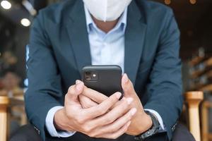 young Businessman in suit wearing surgical face mask and using smartphone, man typing touchscreen mobile phone in office or cafe. Covid-19 Pandemic, technology and New normal concept photo
