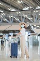 Young female wearing face mask with luggage walking in airport, protection Coronavirus disease infection, Asian woman traveler with hat. Time to travel after vaccine booster dose concept photo