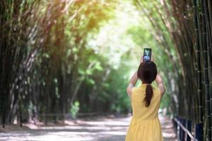 Asian Woman in yellow dress and hat Traveling at green Bamboo Tunnel, Happy traveler taking photo by mobile phone at Chulabhorn wanaram temple in Nakhon Nayok, Thailand