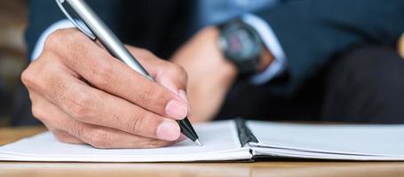 Businessman in suit writing something on notebook in office or cafe, hand of man holding pen with signature on paper report. business concepts photo