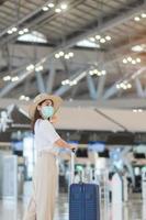 Young female wearing face mask with luggage walking in airport, protection Coronavirus disease infection, Asian woman traveler with hat. Time to travel after vaccine booster dose concept photo