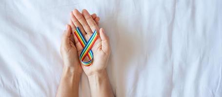 hand showing LGBTQ Rainbow ribbon on white bed background. Support Lesbian, Gay, Bisexual, Transgender, Queer community and Rights concept photo