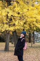 mujer feliz disfruta en el parque al aire libre en la temporada de otoño, viajero asiático con abrigo y sombrero contra el fondo de hojas de ginkgo amarillo foto