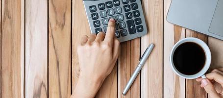 Top view Business woman hand holding coffee cup and calculating with calculator, pen and computer laptop on wood office desk table background. workspace or home office with copy space for text concept photo