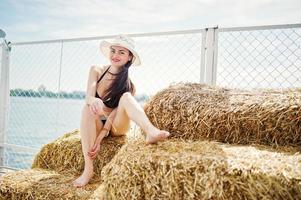 retrato de una chica hermosa en traje de baño bikini negro posando en el fardo de heno con un sombrero junto al lago. foto