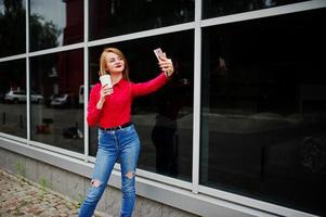 Portrait of a beautiful woman in red blouse and casual jeans taking selfie on mobile phone and holding a cup of coffee outside the huge shopping mall. photo