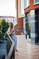 Portrait of a perfect young woman wearing striped overall and yellow sunglasses poses on a balcony of a modern building in a town. photo