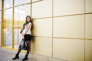 Stylish brunette girl wear on leather jacket and shorts with backpack against golden wall. photo