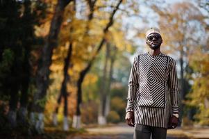 Portrait of stylish black african american man at hat and sunglasses against sunny autumn fall background. Rich people in africa at traditional dress. photo